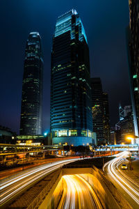 Light trails on road amidst illuminated buildings against sky at night