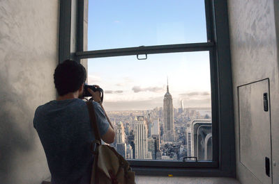 Rear view of man photographing empire state building by window using camera