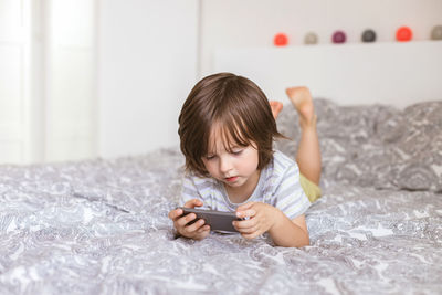 A cute boy, toddler, lies on the bed in the bedroom and plays with the phone, smartphone. problems 