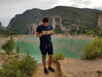 Man reading document while standing at lakeshore against mountains