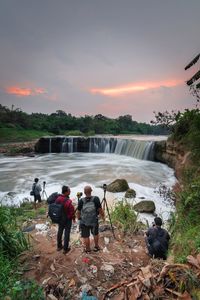 Rear view of people at waterfall against sky during sunset