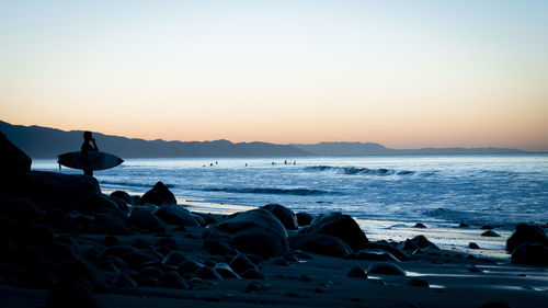 Surfer standing on beach against clear sky during sunset