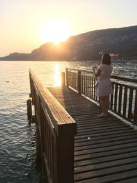 Rear view of woman standing on railing against sea during sunset