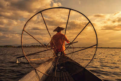 Rear view of man fishing in sea against sunset sky