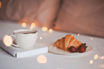 Cup of coffee with open paper book and cake in bed close up over christmas lights. good morning.