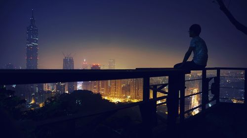 Man looking at taipei 101 while sitting on balcony railing in city at night