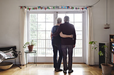 Full length rear view of retired couple looking through window at home