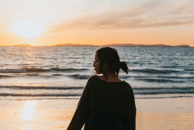 Side view of young woman standing at beach during sunset