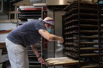 Side view of female cook adding flour on raw baguettes while preparing bread for baking in kitchen