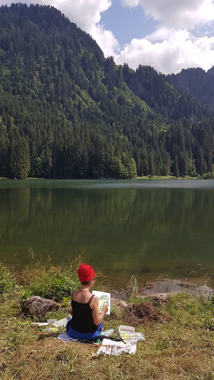 WOMAN SITTING ON LAKE AGAINST MOUNTAINS