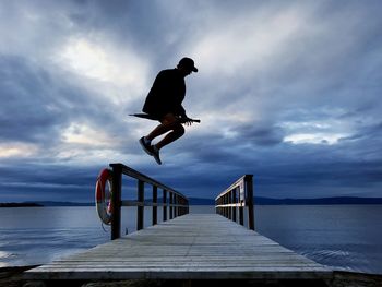 Man jumping over jetty against cloudy sky