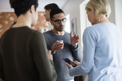 Man with glass gesturing while talking to woman holding phone at social gathering