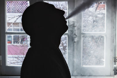 Silhouette man exhaling smoke against window