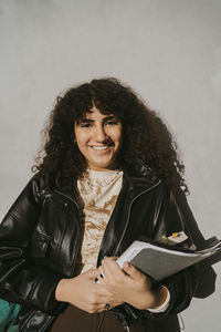 Portrait of smiling young woman wearing leather jacket holding book leaning on gray wall