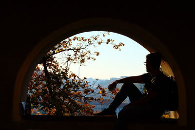 Silhouette man sitting by window against sky