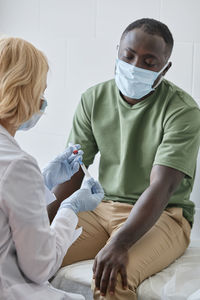 Patient sitting on bed with doctor preparing vaccine syringe in clinic