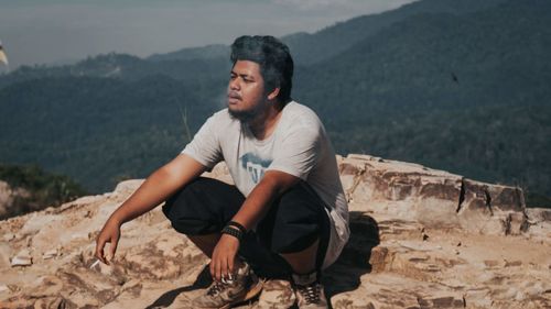 Young man looking away and smoking while sitting on rock against mountains