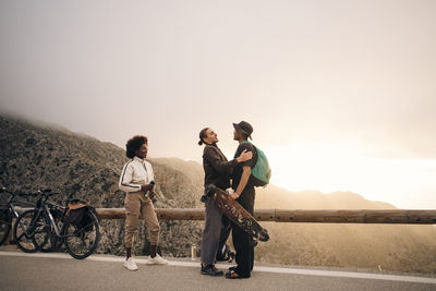 Young male and female friends greeting each other while standing near wooden guardrail on road against sky