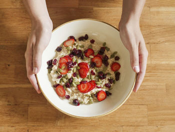 Woman's hands hold a bowl of oatmeal with strawberries on a wooden table. view from above.