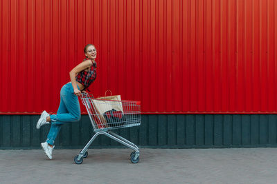 Happy girl with shopping cart on red wall shop background. young woman pushing a shopping cart 