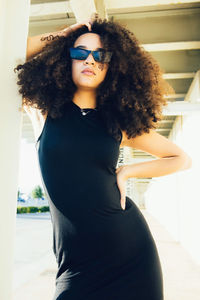 Cool young ethnic woman with afro curls wearing black dress and sunglasses standing near stone column with hand on waist under roof near building in sunny day in street