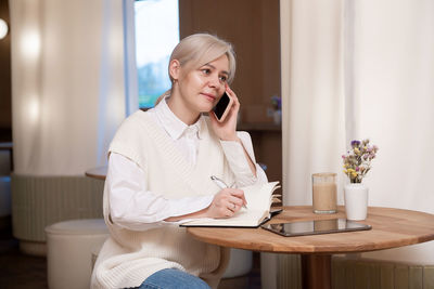 A cute girl is sitting in a cafe and talking on the phone and making notes in a diary