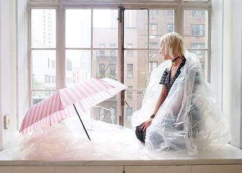 Woman wrapped in plastic by umbrella looking through window at home