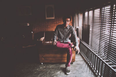 Full length portrait of young man sitting by window blinds at home