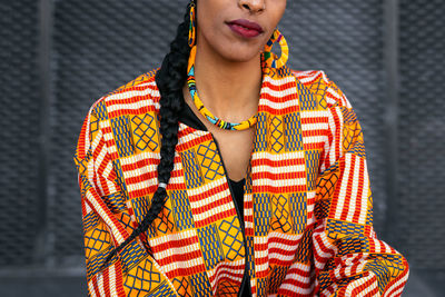 Cropped unrecognizable ethnic woman in colorful shirt with braid near gray wall in daytime on city street