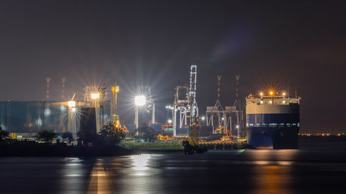 Illuminated factory by sea against sky at night