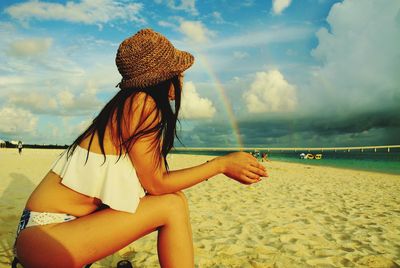 Side view of woman in bikini sitting at beach against sky