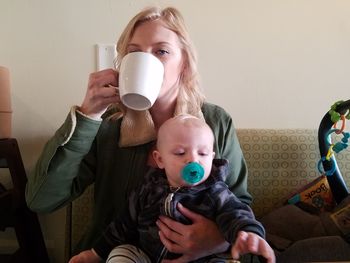 Portrait of mother drinking coffee while carrying son at home