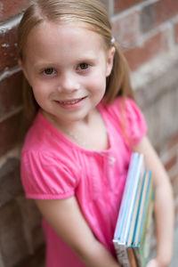 Portrait of smiling girl holding books by brick wall