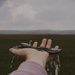 Cropped hand holding wet mobile phone with toy animal against sky