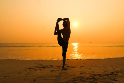 Full length of silhouette woman practicing yoga at beach against dramatic sky during sunset