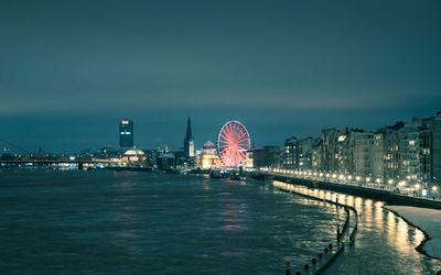 Illuminated ferris wheel by river against sky in city at night