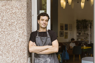 Portrait of smiling male owner with arms crossed leaning on wall near store doorway