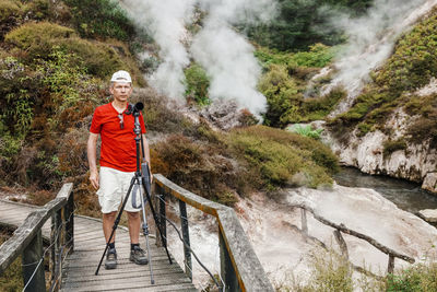 Senior man with camera tripod standing on footbridge by stream