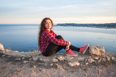 Portrait of smiling woman sitting on rock by sea against sky