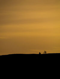 Silhouette people riding horse on field against orange sky