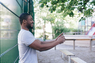 Side view of smiling man taking selfie while standing against fence