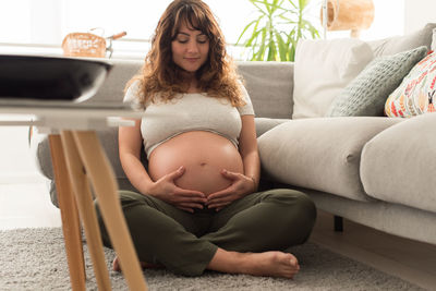 Pregnant mid adult woman touching belly while sitting by sofa on rug at home