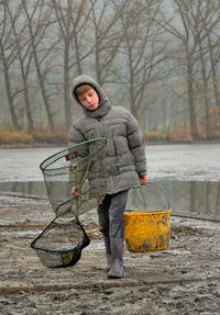 Boy carrying bucket and fishing net while walking on field by lake