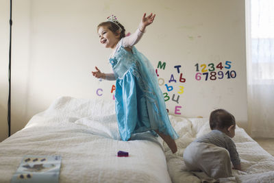 Happy girl wearing princess costume jumping on bed with baby brother negative space