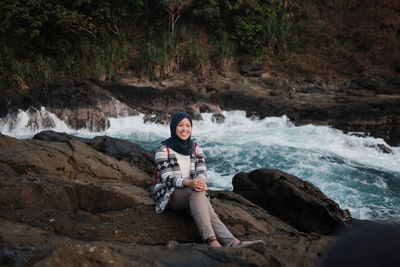 Smiling woman wearing hijab sitting on rock by river