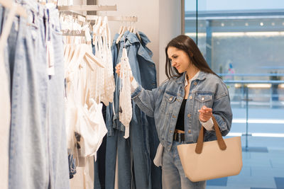 Young woman stands in a fashion store, carefully choosing clothing items to buy. she browses through