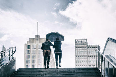 Rear view of women with umbrella at staircase against sky during rainy season