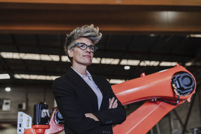 Thoughtful businesswoman with arms crossed standing in front of robotic arm