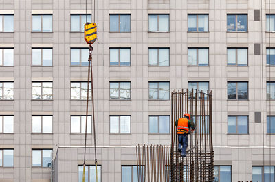 A worker in a bright orange vest assembles a metal frame made of steel reinforcement at the height.