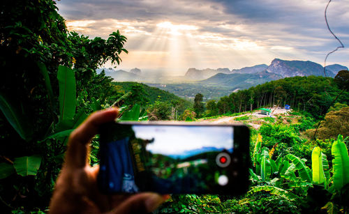 Cropped hand of man photographing through smart phone against sky